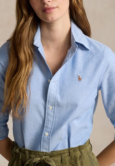 Noor naine, kellel on pikk pruun juuksed, seljas hele sinine nööpidega särk väikese tikandiga hobuse logoga ja oliivirohelised püksid.