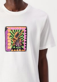 White cotton t-shirt featuring a colorful graphic of a frog playing guitar with pink and black accents, framed by a geometric background.