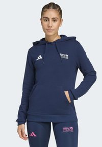 Kvinde iført en marineblå BMW Berlin Marathon hoodie og leggings med logoer, der står med den ene hånd i hoodie-lommen.