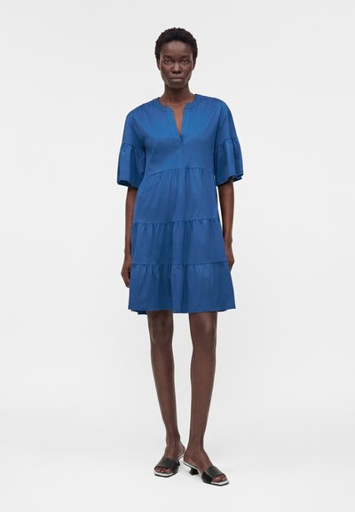 Γυναίκα όρθια που φοράει μπλε φόρεμα με βολάν μέχρι το γόνατο, με μανίκια μέχρι τον αγκώνα και μαύρα ανοιχτά ψηλοτάκουνα σανδάλια σε λευκό φόντο.