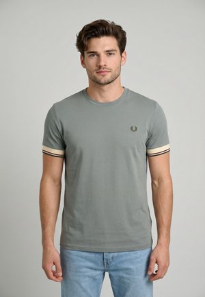 Giovane uomo con capelli castani corti che indossa una t-shirt verde a maniche corte con polsini a righe e jeans azzurri chiari su uno sfondo semplice.