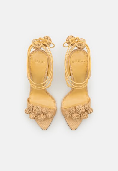 Yellow high-heeled sandals with a pointed toe, featuring woven accents and rounded pom-poms on the front, showing a textured fabric design.