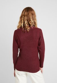 Femme aux cheveux ondulés châtain clair portant un pull bordeaux à manches longues et un pantalon blanc, debout face à un fond uni gris clair.