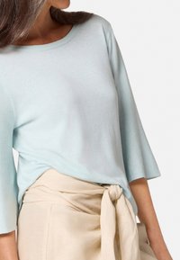  Lichtblauwe, kortgebalokte trui met ronde halslijn; zachte textuur; gecombineerd met beige, hooggehaalde broek met strik om de taille.