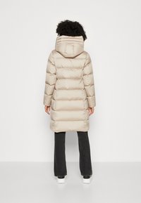 Abrigo acolchado beige con capucha, con secciones acolchadas y diseño de longitud media. Combinado con pantalones negros y zapatos blancos.