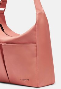 Różowa syntetyczna torba na ramię o gładkiej teksturze, z zamkiem błyskawicznym i przednią kieszenią. Logo widoczne w pobliżu podstawy.