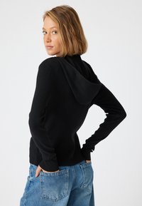 Svart ribbad stickad hoodie med avslappnad passform, har en mjuk huva och långa ärmar, kombinerad med ljusblå denimjeans.