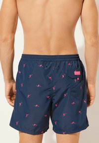 Bañador de color navy con un patrón de flamencos rosas, cinturilla elástica y un bolsillo trasero con un parche de la marca. Tela suave, longitud a mitad del muslo.