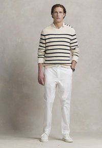 Gestreifter Strickpullover in Creme und Marine, kombiniert mit weißen Hosen und weißen Sneakern. Auffälliges Knopfdetail an der Schulter.
