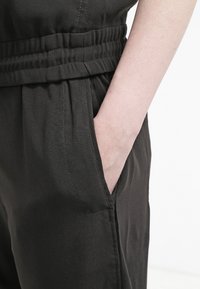 Czarne materiałowe spodnie z teksturowanym pasem i bocznymi kieszeniami, z jedną ręką spoczywającą w kieszeni. Prosty i funkcjonalny design.