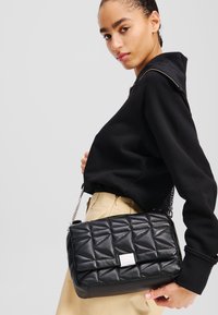 Borsa a tracolla nera trapuntata in pelle, con chiusura a patta, tracolla a catena e motivi geometrici sulla superficie.