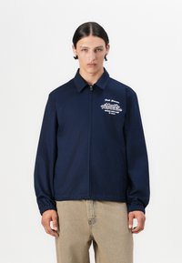 Navy zip-up jacket with relaxed fit, elastic cuffs, chest embroidery, and side pockets. Worn over beige pants. Smooth fabric texture.