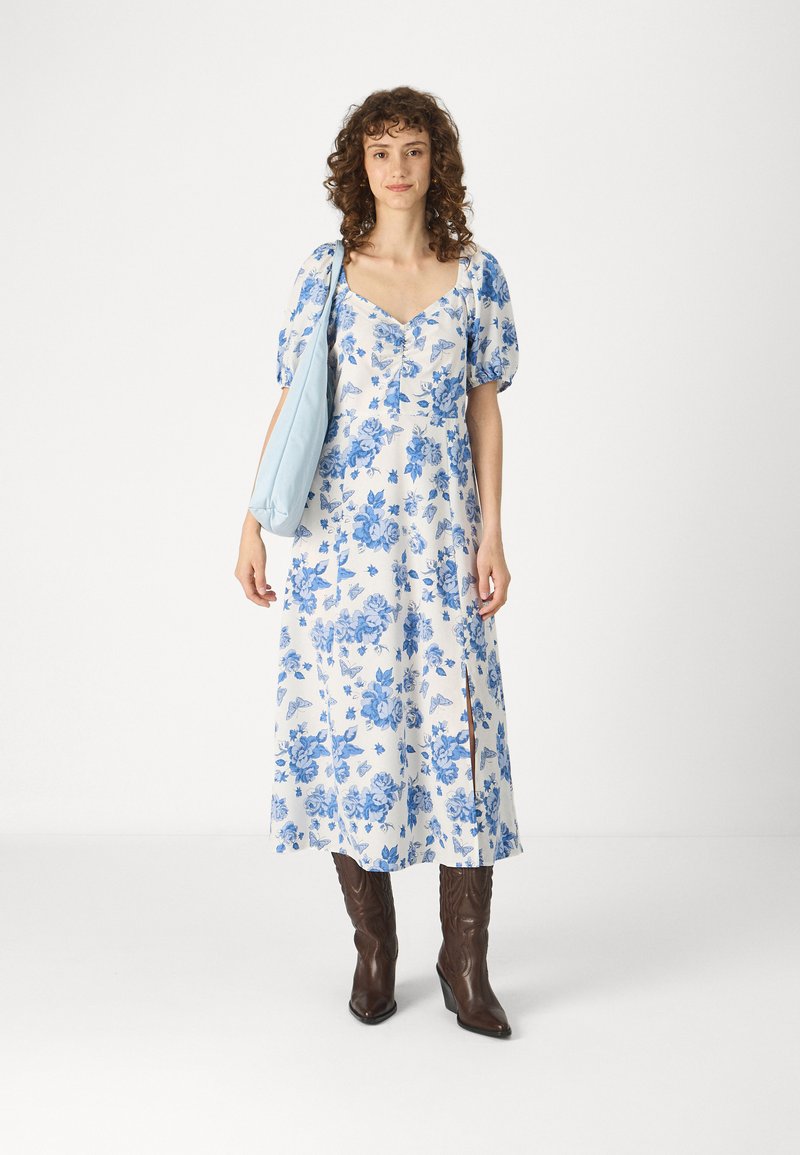 Robe fleurie blanche avec des motifs bleus, manches bouffantes, décolleté en V et deux fentes latérales. Associée à des bottes à talons brunes et un sac bleu clair.