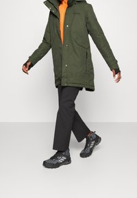 Parka verde con cappuccio, dotata di due tasche frontali e una cerniera. Indossata sopra una maglietta arancione, abbinata a pantaloni neri e scarpe grigie.