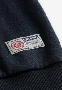 Tmavomodrý textil s bielou obdĺžnikovou vyšitou etiketou, na ktorej je text "SUPERDRY" a "Vintage Athletic Outfitters" a červeným kruhovým logom.
