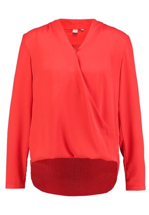 Woman wearing a bright red long-sleeve wrap blouse paired with dark gray high-waisted jeans against a plain background.