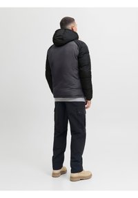 Mężczyzna zwrócony tyłem, ubrany w czarno-szarą pikowaną kurtkę z kapturem, czarne spodnie typu cargo oraz beżowe buty na jednolitym białym tle.