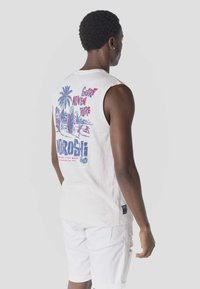 Hombre de espaldas con camiseta sin mangas blanca con estampado colorido de surf y playa, combinada con pantalones cortos blancos sobre fondo liso.