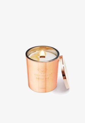 Candela in un barattolo metallico oro rosa con bordo nero, con fiamma visibile e due cristalli di citrino incastonati nella cera. Coperchio accanto.