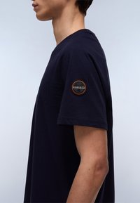 Navy blue cotton t-shirt with a round neckline and short sleeves. Features a textured logo and a circular patch on the left sleeve.