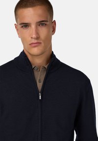 Tamsiai mėlynas džemperis su užtrauktuku, aukštu apykakle, aptemptas dizainas ir tekstūrinis trikotažas, dėvimas ant šviesiai smėlio spalvos marškinių su sagomis.