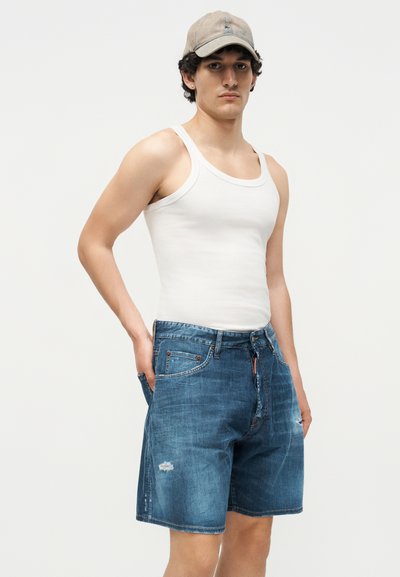 Άνδρας που φοράει μπεζ καπέλο, λευκή φανέλα και μπλε ξεβαμμένο τζιν σορτς με τα χέρια στις τσέπες, σε απλό φόντο.