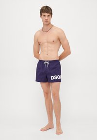 En ung mann uten skjorte som har på seg lilla badshorts med hvit "DSQU" tekst, står barbeint med hendene bak ryggen på en ensfarget bakgrunn.