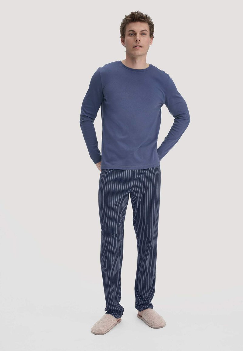 Marineblaues Langarmoberteil, kombiniert mit marineblauen gestreiften Hosen. Weicher Stoff, lockere Passform und hellgraue Hausschuhe ergänzen den lässigen Look.