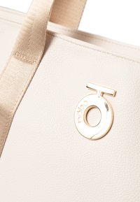 Crème faux leren totebag met een textuur oppervlak, voorzien van een goudkleurig cirkelvormig logo-accent en beige stoffen hengsels.