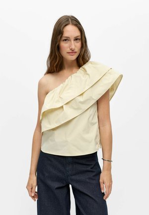 Femme aux longs cheveux bruns portant un haut crème à une épaule volantée et un pantalon bleu foncé, debout sur un fond blanc.
