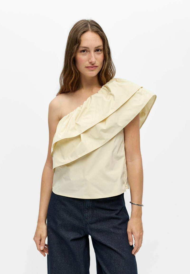 Femme aux longs cheveux bruns portant un haut crème à une épaule volantée et un pantalon bleu foncé, debout sur un fond blanc.