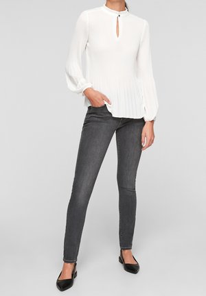 Frau trägt eine weiße, langärmelige, plissierte Bluse mit Schlitz am Ausschnitt, dunkelgraue Skinny-Jeans und schwarze spitze Ballerinas.