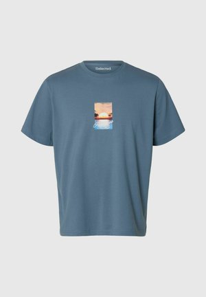 T-shirt bleu à manches courtes avec un graphisme coucher de soleil et océan sur la poitrine et le mot « Paradise » en cursive sous l'image.