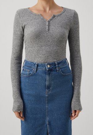 Femme portant un haut gris côtelé ajusté à manches longues avec des boutons et une jupe en jean bleue taille haute avec une fermeture éclair devant et des poches.