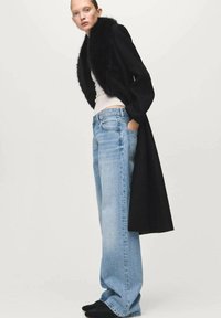 Lange zwarte jas met een faux fur kraag, gedragen over een strak wit topje en losse lichtblauwe spijkerbroeken. Zwarte schoenen maken de outfit compleet.