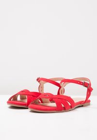 Sandales en daim rouge avec semelle plate, bretelles croisées et boucle réglable à la cheville. Design à bout ouvert avec motifs découpés pour une meilleure respirabilité.