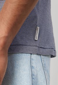 Marineblauwe en witte gestreepte t-shirt met een label aan de zijkant. De stof is zacht, gecombineerd met lichtblauwe denimshorts op de achtergrond.