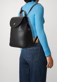 Czarny skórzany plecak o zaokrąglonym kształcie, z klapą na zagięcie i złotymi akcentami. Noszony na plecach, w połączeniu z niebieską koszulą z długim rękawem i dżinsami.