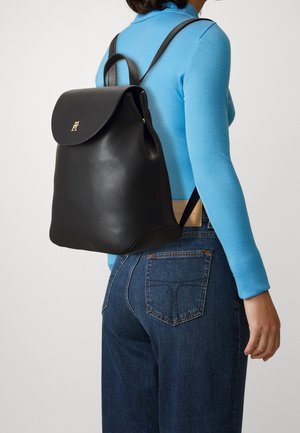 Mochila de cuero negro con forma redondeada, tapa plegable y herrajes dorados. Llevada en la espalda, combinada con una camisa de manga larga azul y vaqueros.