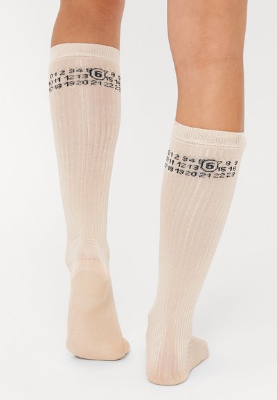 Chaussettes beiges jusqu'aux genoux avec texture côtelée. Imprimé numérique noir sur le bandeau supérieur. Matériau doux, design ajusté avec un orteil sans couture.