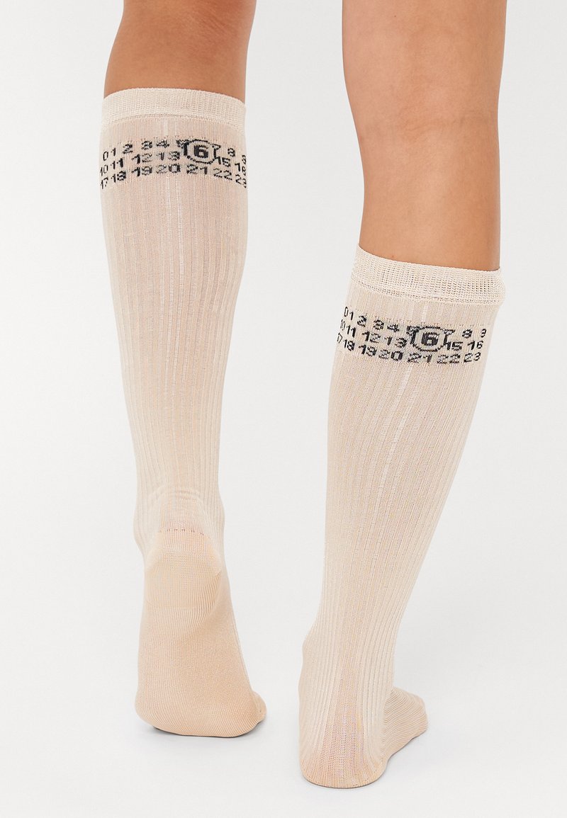 Chaussettes beiges jusqu'aux genoux avec texture côtelée. Imprimé numérique noir sur le bandeau supérieur. Matériau doux, design ajusté avec un orteil sans couture.