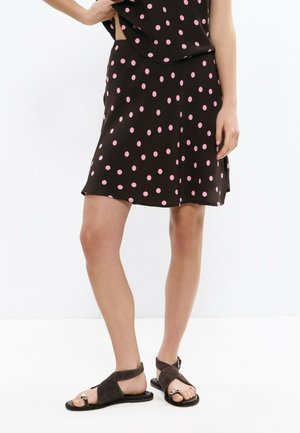 Mujer con falda negra con lunares rosas y sandalias marrón oscuro de punta abierta, de pie con una mano en la cadera contra un fondo blanco.