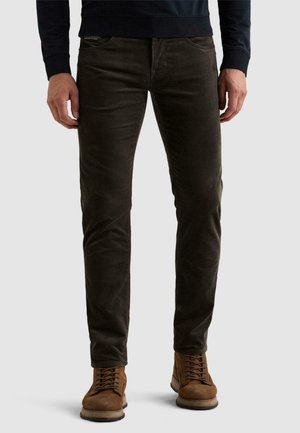 Homme portant un pantalon en velours côtelé marron foncé ajusté et des bottes marron à lacets, debout devant un fond clair uni.