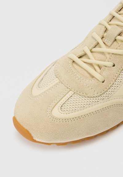 Μπεζ αθλητικό παπούτσι με υλικά από σουέντ και δίχτυ, στρογγυλεμένη μύτη και υφασμάτινες λεπτομέρειες. Κρεμ κορδόνια και εξωτερική σόλα από καουτσούκ χρώματος γκαμ ολοκληρώνουν το σχέδιο.