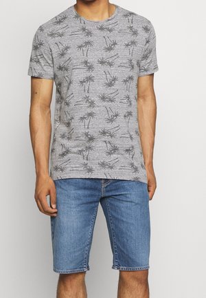Mand iført en grå t-shirt med sorte palmeprint og blå denim knælange shorts mod en ensfarvet baggrund.