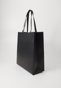 Sac fourre-tout noir avec texture en cuir lisse, forme rectangulaire, deux longues poignées et détail de poche avant. Design minimaliste et spacieux.