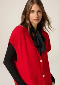 Gilet rosso lavorato a maglia con maniche ampie, chiusura con bottoni e due tasche frontali. Indossato sopra una maglia nera a maniche lunghe con un grande fiocco decorativo.