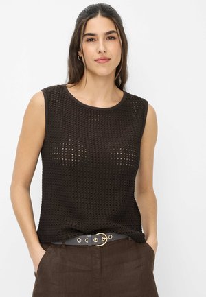 Jeune femme aux cheveux foncés portant un haut sans manches en tricot noir et un pantalon marron, debout les mains dans les poches devant un fond blanc.