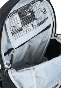 Interior de una mochila que muestra una etiqueta con las instrucciones de emergencia, incluyendo texto e íconos sobre primeros auxilios, llamada de emergencia y señales de seguridad.
