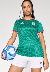 Camisa de futebol verde com um padrão texturizado, detalhes vermelhos nas mangas e o logo da Adidas; acompanhada de calções brancos e uma bola azul.